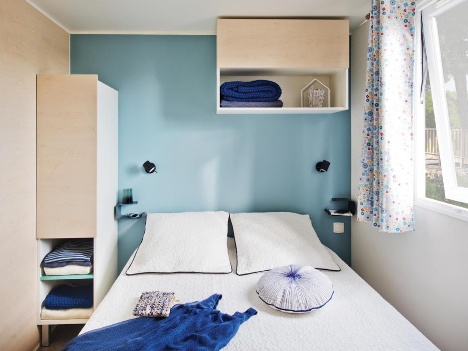 Sie sehen ein Doppelbett in einem Mobile Home beim JUFA Vulkan Thermen-Resort mit großem Fenster. JUFA Hotels bietet erholsamen Thermenspass für die ganze Familie.