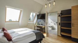 Sie sehen ein Doppelbett, Schreibtisch, Schrank und Badezimmer im Doppelzimmer Deluxe im JUFA Hotel Bregenz am Bodensee. Der Ort für einen unvergesslichen Wander-, Rad- und Kultururlaub für Familien, Freunde und Paare.