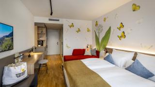 Sie sehen ein Doppelzimmer superior für bis zu 3 Personen im JUFA Hotel Bad Radkersburg mit komfortablem Boxpringbett, gemütlichem Sofa, modernem Möbliliar, kreativer Wanddeko und einer Badetasche am Zimmer