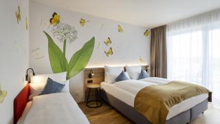 Sie sehen ein Doppelzimmer superior für 3 Personen im JUFA Hotel Bad Radkersburg mit Balkon, komfortablem Boxpringbett, gemütlichem Sofa, und kreativer Wanddeko.
