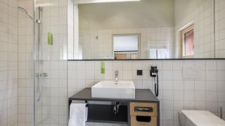 Sie sehen eine barrierefreie Dusche, ein Waschbecken, eine Toilette und einen Spiegel im Badezimmer eines Doppelzimmer superior im JUFA Hotel Bregenz am Bodensee. Der Ort für einen unvergesslichen Wander-, Rad- und Kultururlaub für Familien, Freunde und Paare.