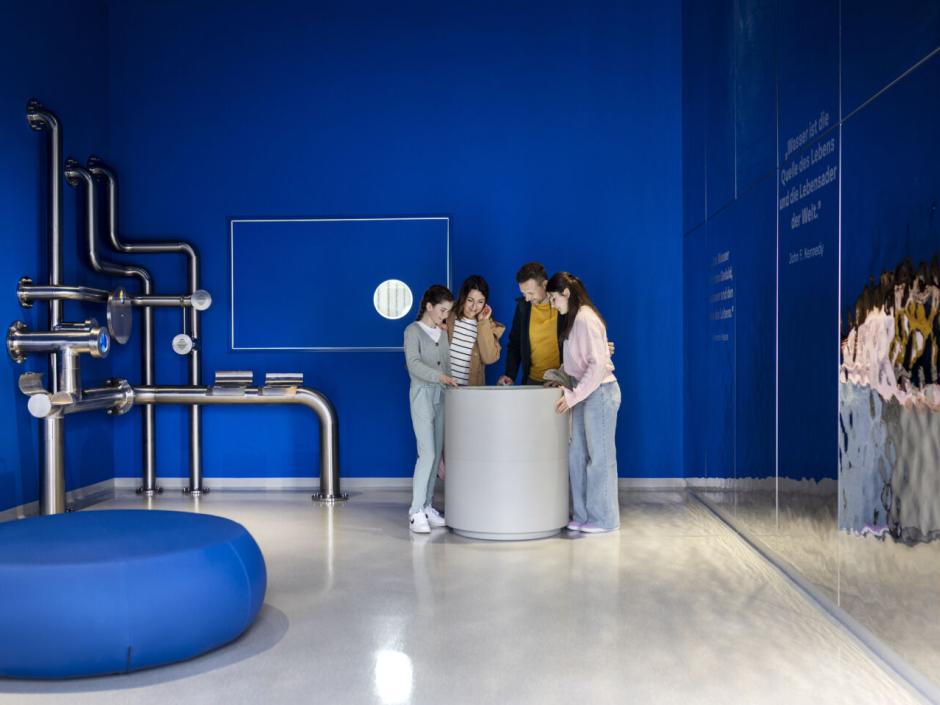 Sie sehen eine vierköpfige Familie an einem interaktiven Ausstellungselement im Raum "Quell des Lebens" im JUFA Hotel Bad Radkersburg.