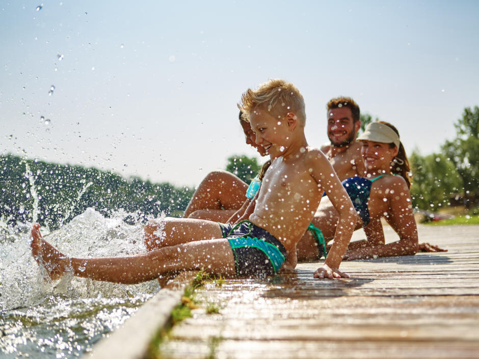 Sie sehen eine Familie beim Baden am Steg. JUFA Hotels bietet tollen Sommerurlaub an schönen Seen für die ganze Familie.