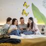 Sie sehen eine lachende vierköpfige Familie gemütlich auf einem Bett sitzend beim gemeinsamen Entdecken von digitalen Inhalten auf einem Tablet im JUFA Hotel Bad Radkersburg