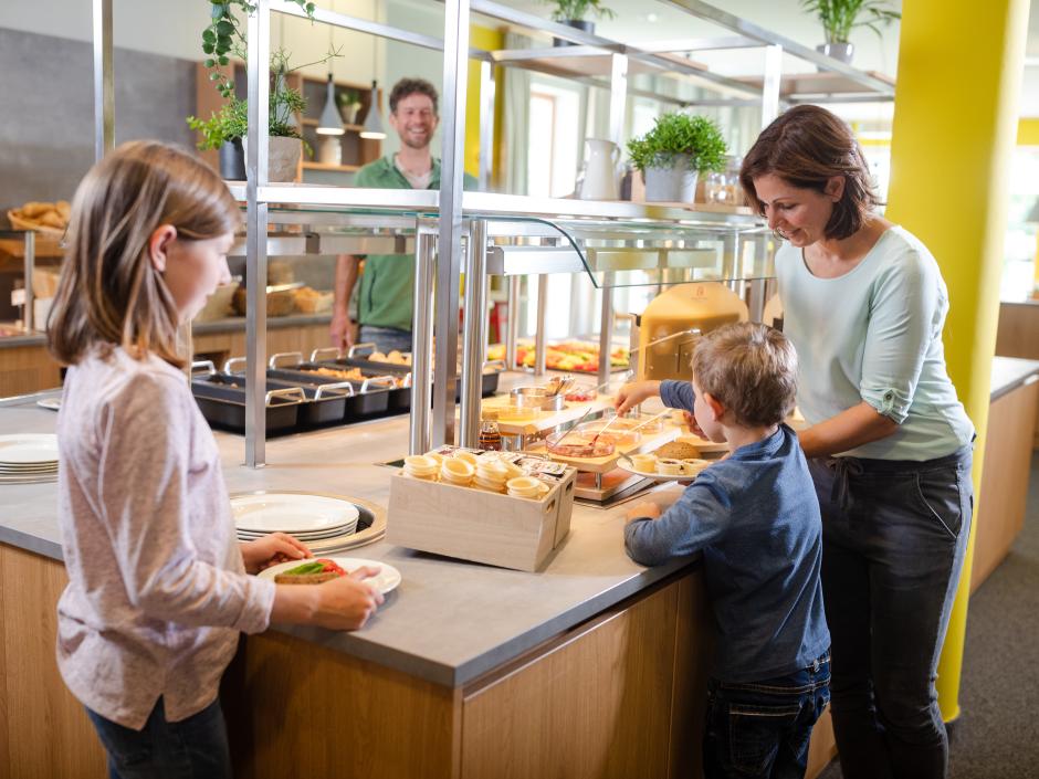 Sie sehen eine Familie am Frühstücksbuffet im JUFA Kempten mit herzhafter oder süßer Auswahl. JUFA Hotels bietet kinderfreundlichen und erlebnisreichen Urlaub für die ganze Familie.