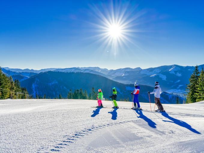 Sie sehen eine Familie beim Skifahren mit Sonne und Bergen auf der Mariazeller Bürgeralpe. JUFA Hotels bietet erholsamen Familienurlaub und einen unvergesslichen Winterurlaub.