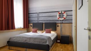 Sie sehen ein Doppelbett vor einer Fototapete und Rettungsring-Dekoration im Doppelzimmer oder Family & Friends 3 plus Zimmer im JUFA Hotel Bregenz am Bodensee. Der Ort für einen unvergesslichen Wander-, Rad- und Kultururlaub für Familien, Freunde und Paare.