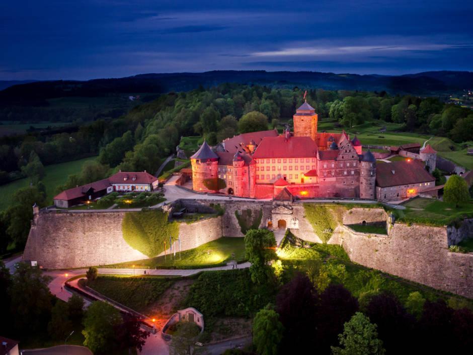 Sie sehen die Festung Rosenberg in Kronach mit Lichtern am Abend. JUFA Hotels bietet kinderfreundlichen und erlebnisreichen Urlaub für die ganze Familie.