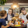 Sie sehen eine Familie am Frühstückstisch mit Mama, Papa, Tochter und Sohn. JUFA Hotels bietet erholsamen Familienurlaub und einen unvergesslichen Winter- und Wanderurlaub.