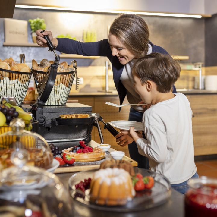Sie sehen wie die Mutter ihrem Sohn hilft Waffeln beim Frühstücksbuffet zuzubereiten. JUFA Hotels bietet erholsamen Familienurlaub und einen unvergesslichen Winter- und Wanderurlaub.
