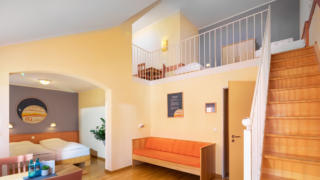 Sie sehen ein Galeriezimmer mit Doppelbett und Sitzbank für sechs Personen im JUFA Hotel Nördlingen. Der Ort für kinderfreundlichen und erlebnisreichen Urlaub für die ganze Familie.