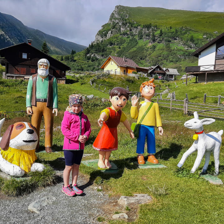 Sie sehen ein Mädchen, das vor bunten Heidi Figuren auf der Alm steht. JUFA Hotels bietet erholsamen Ski- und Wanderurlaub für Familien in Kärnten.