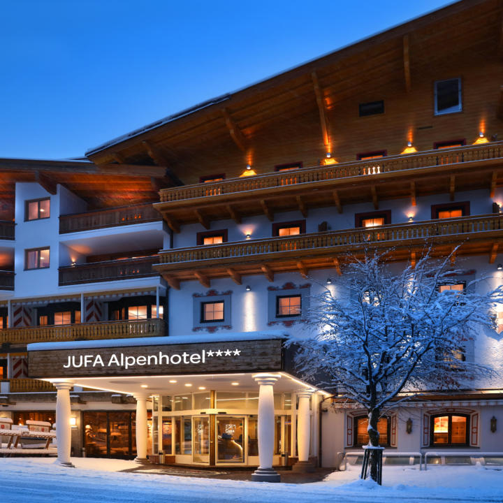 Sie sehen eine Außenansicht vom JUFA Alpenhotel Saalbach**** am Abend im Winter. JUFA Hotels bietet erholsamen Familienurlaub und einen unvergesslichen Winterurlaub.