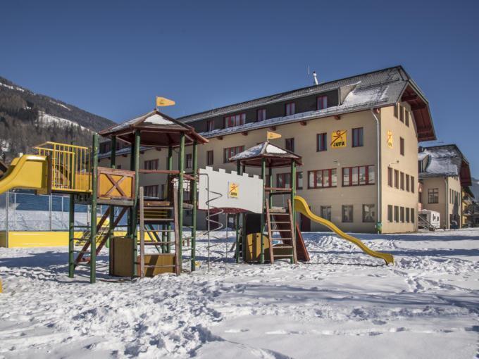 Hotelansicht vom JUFA Hotel Lungau mit Kletterburg vor blauem Himmel im Winter. JUFA Hotels bieten erholsamen Familienurlaub und einen unvergesslichen Winterurlaub.