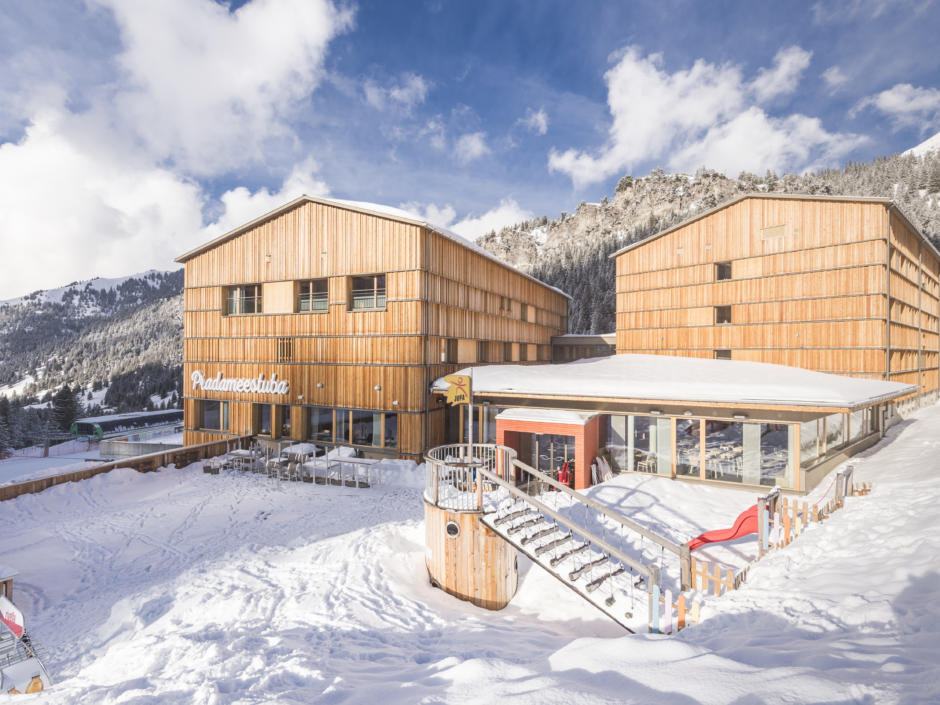 Sie sehen eine Außenansicht vom JUFA Hotel Malbun – Alpin-Resort*** mit Spielplatz im Winter. JUFA Hotels bietet erholsamen Familienurlaub und einen unvergesslichen Winterurlaub.