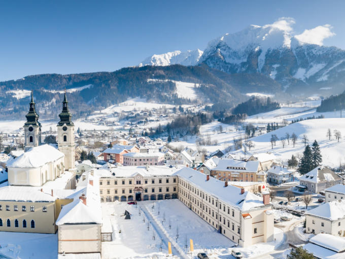 Sie sehen eine Außenansicht vom JUFA Hotel Pyhrn-Priel mit Bergen im Winter. JUFA Hotels bietet erholsamen Familienurlaub und einen unvergesslichen Winterurlaub.