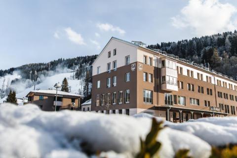 Sie sehen eine Außenansicht vom JUFA Hotel Schladming mit Schnee im Winter. JUFA Hotels bietet erholsamen Familienurlaub und einen unvergesslichen Winterurlaub.