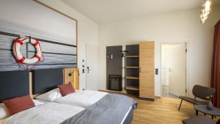Sie sehen ein Doppelbett, einen Beistelltich mit zwei Stühlen sowie einen Schrank und einen Spiegel im Doppelzimmer deluxe im JUFA Hotel Bregenz am Bodensee. Der Ort für einen unvergesslichen Wander-, Rad- und Kultururlaub für Familien, Freunde und Paare.