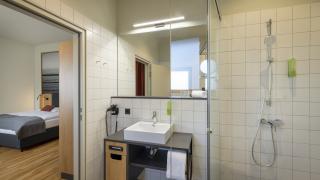 Sie sehen ein Badezimmer mit Waschbecken, Spiegel, Föhn und Dusche im Doppelzimmer deluxe im JUFA Hotel Bregenz am Bodensee. Der Ort für einen unvergesslichen Wander-, Rad- und Kultururlaub für Familien, Freunde und Paare.