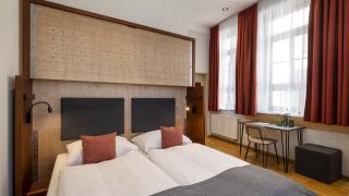 Sie sehen ein Doppelbett sowie einen Schreibtisch im Doppelzimmer im JUFA Hotel Bregenz am Bodensee. Der Ort für einen unvergesslichen Wander-, Rad- und Kultururlaub für Familien, Freunde und Paare.