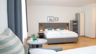 Sie sehen ein Bild vom Doppelbett im Family & Friends Zimmer im JUFA Hotel Garni Stubenberg in der Oststeiermark mit Sitzecke und Kasten.