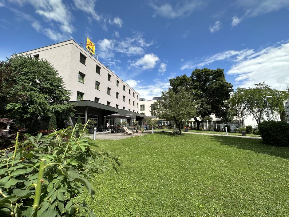 Sie sehen das JUFA Hotel Graz City in Schrägansicht von außen, davor Büsche, Bäume und eine grüne Wiese. Der Ort für einen erlebnisreichen Städtetrip für die ganze Familie und der ideale Platz für Ihr Seminar.