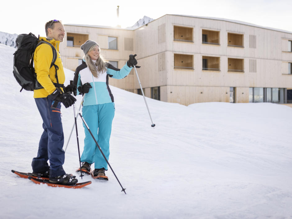 Sie sehen ein Pärchen beim Schneeschuh Wandern. Im Hintergrund ist das JUFA Hotel Laterns zu sehen. Der Ort für erholsamen Familienurlaub und einen unvergesslichen Winter- und Wanderurlaub.