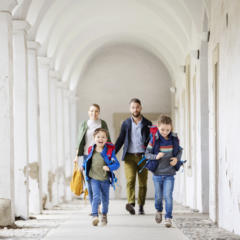 Sie sehen eine Familie von vorne mit Muter, Vater, Tochter, Sohn. Die Kinder laufen auf die Kamera zu, umgeben von den Klosterbögen vom JUFA Klosterhotel Judenburg. Der Ort für erholsamen Familienurlaub und einen unvergesslichen Winter- und Wanderurlaub.