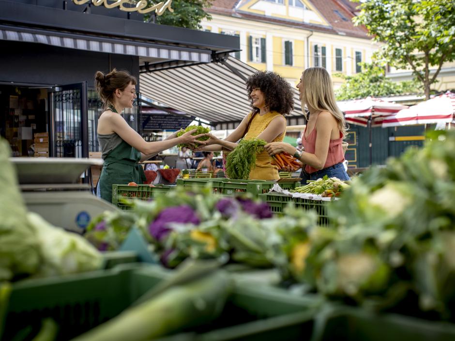 Sie sehen zwei junge Frauen, die am Kaiser-Josef-Markt in Graz bei einer Frau Gemüse kaufen.