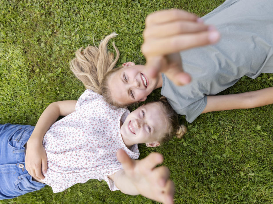 Sie sehen zwei Mädchen im Gras liegen, die mit ihren Fingerin in die Höhe zeigen und lächeln.