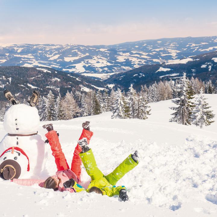 Sie sehen zwei Kinder, die im Schnee spielen neben einen selbstgebauten Schneemann am Klippitztörl
