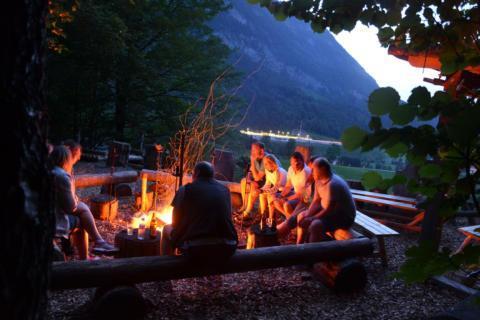 Eine Gruppe Erwachsener sitzt in Pyhrn-Priel im Wald am Lagerfeuer und grillt. JUFA Hotels bietet den Ort für erfolgreiche und kreative Seminare in abwechslungsreichen Regionen.