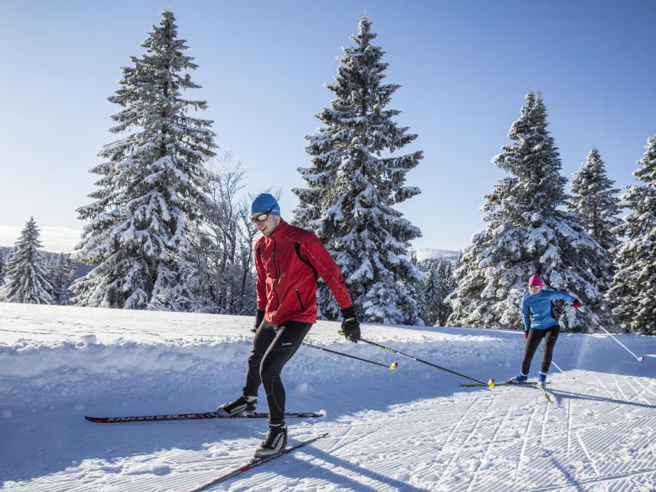 Sie sehen 2 Personen beim Langlaufen auf der Loipe Herzogenhorn mit winterlicher Landschaft im Hintergrund.