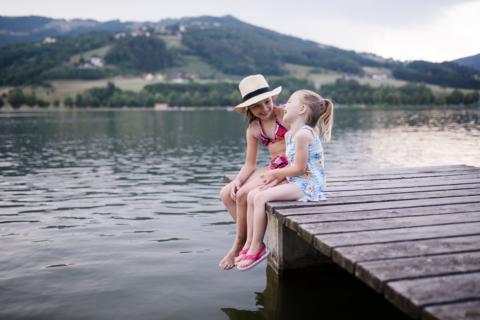 Sie sehen zwei Mädchen auf einem Steg am Stubenbergsee in Badekleidung im Sommer. JUFA Hotels bietet tollen Sommerurlaub an schönen Seen für die ganze Familie.