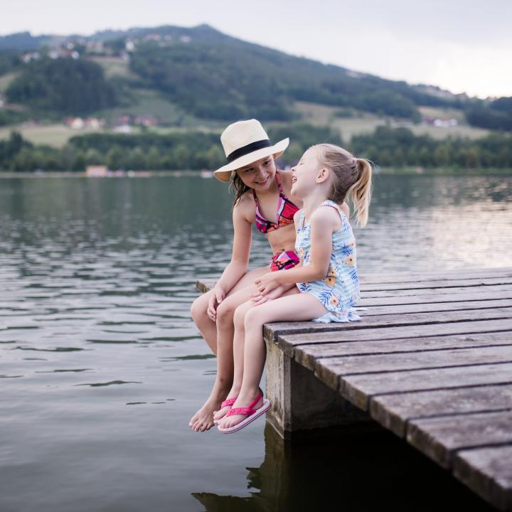 Sie sehen zwei Mädchen auf einem Steg am Stubenbergsee in Badekleidung im Sommer. JUFA Hotels bietet tollen Sommerurlaub an schönen Seen für die ganze Familie.