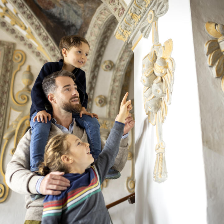 Sie sehen einen Vater mit seinen beiden Kindern. Die Tochter und der Vater zeigen auf das Wandrelief aus Stein im Museum Murtal. JUFA Hotels bietet kinderfreundlichen und erlebnisreichen Urlaub für die ganze Familie.