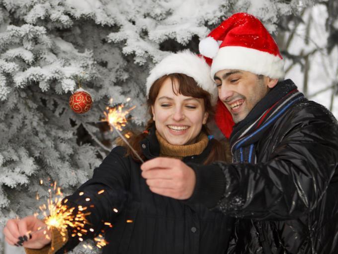 Ein Paar mit Weihnachtsmannmützen steht vor verschneiten Bäumen und hält Spritzkerzen. JUFA Hotels bietet erholsamen Familienurlaub und einen unvergesslichen Winterurlaub.