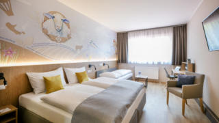 Sie sehen einen Raumblick mit Bett im Doppelzimmer im JUFA Hotel Weiz. Der Ort für kinderfreundlichen und erlebnisreichen Urlaub für die ganze Familie.