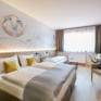 Sie sehen einen Raumblick mit Bett im Doppelzimmer im JUFA Hotel Weiz. Der Ort für kinderfreundlichen und erlebnisreichen Urlaub für die ganze Familie.