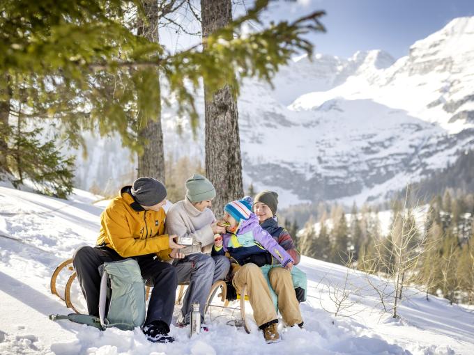 Sie sehen eine Familie mit Vater, Mutter, Tochter und Sohn mit einer Jausenbox und Thermosflasche auf einer Rodel in einer verschneiten, sonnigen Winterlandschaft mit Bergen im Hintergrund.