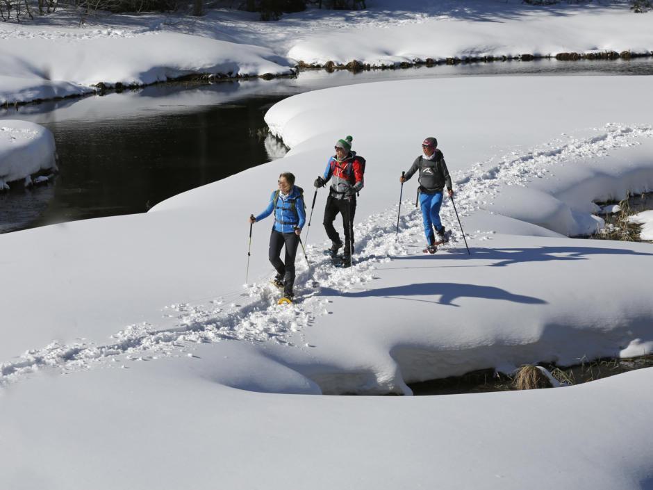 Sie sehen Schneeschuhwanderer an einem Fluss im Murtal im Winter wandern. JUFA Hotels bietet erholsamen Familienurlaub und einen unvergesslichen Winterurlaub.