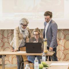 Zwei Seminarteilnehmer und die Seminarleiterin versammeln sich um einen Laptop im JUFA Hotel Wien City. Der Ort für erlebnisreichen Städtetrip für die ganze Familie und der ideale Platz für Ihr Seminar.