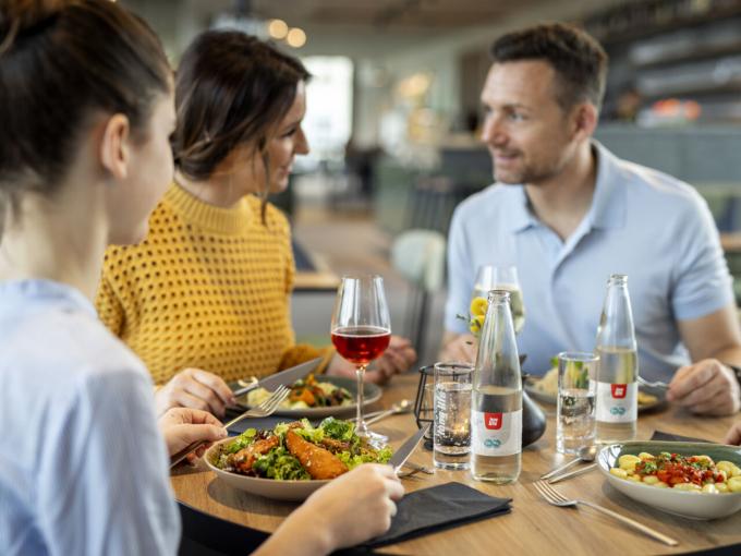 Sie sehen eine vierköpfige Familie beim gemütlichen Mittag- und Abendessen mit regionalen Speisen und Getränken im Café & Restaurant Wandelhalle im JUFA Hotel Bad Radkersburg