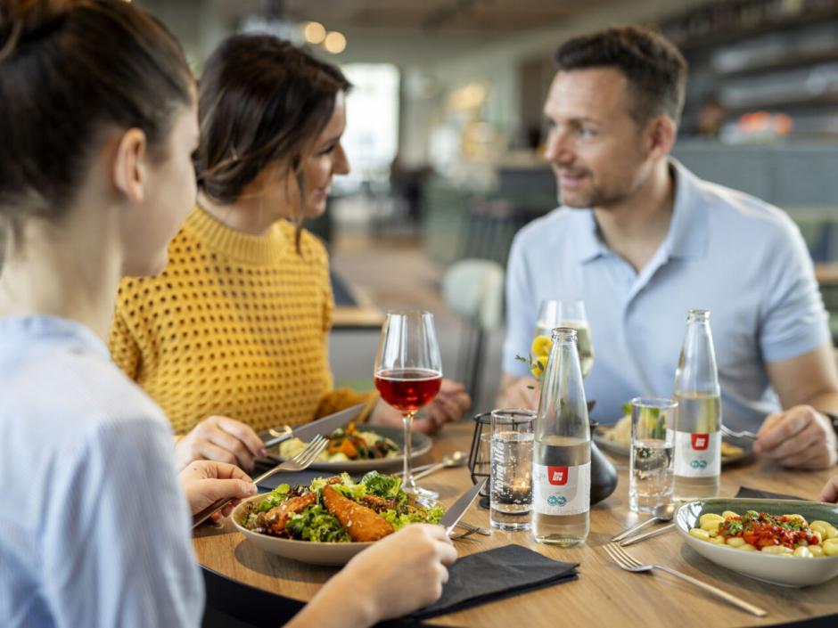 Sie sehen eine vierköpfige Familie beim gemütlichen Mittag- und Abendessen mit regionalen Speisen und Getränken im Café & Restaurant Wandelhalle im JUFA Hotel Bad Radkersburg