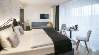 Sie sehen ein Doppelbett und eine Couch in einer Suite des JUFA Hotels Hamburg HafenCity. Der Ort für erlebnisreichen Städtetrip für die ganze Familie und der ideale Platz für Ihr Seminar.