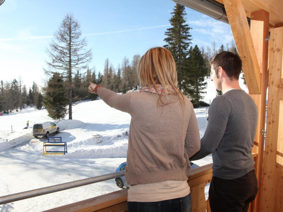 Sie sehen zwei Menschen auf der Terrasse eines Hotelzimmers mit Blick auf eine Winterlandschaft im JUFA Hotel Nockberge - Almerlebnis. Der Ort für erholsamen Familienurlaub und einen unvergesslichen Winter- und Wanderurlaub.