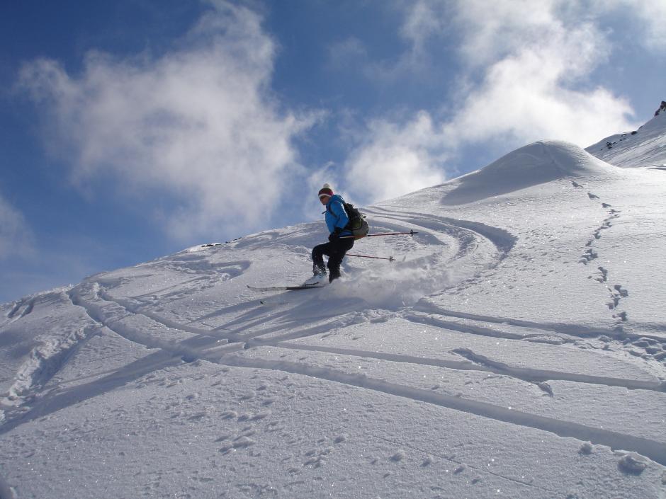 Sie sehen einen Skitourengeher bei der Abfahrt über einen verschneiten Hang bei Sonne und blauem Himmel.