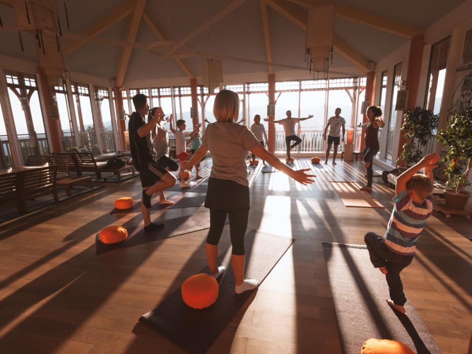 Eine Yogagruppe macht Yoga im Sonnengrußraum und durch die tiefstehende Sonnen werfen die Teilnehmer lange Schatten.JUFA Hotels, der Ort für erholsamen Familienurlaub und einen unvergesslichen Winter- und Wanderurlaub.