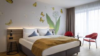 Sie sehen ein Doppelzimmer für zwei Personen im JUFA Hotel Bad Radkersburg mit Balkon, komfortablem Boxpringbett, modernem Möbiliar, gemütlicher Sitzecke mit Tisch und kreativer Wanddeko.