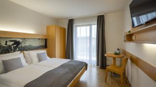 Sie sehen ein Doppelbett mit Zierpolster und Bettschal, eine Sitzecke, einen Kasten sowie den Ausgang zum Balkon oder zur Terrasse im JUFA Hotel Schilcherland*** in der Steiermark.